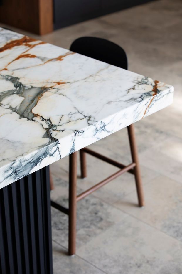 Close-up of a dramatic white natural stone tabletop with prominent grey, black, and rust-coloured veining. The thick slab has a mitred or solid-looking edge and is paired with a simple black curved-back stool with a brown timber or metal frame. The setting has large-format pale stone floor tiles and dark architectural joinery in the background, creating a refined contemporary interior product-detail view.
