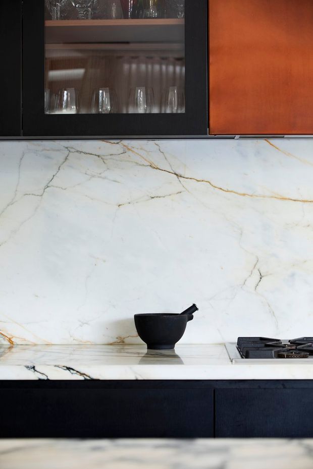 Close-up view of a refined kitchen with dark cabinetry, black-framed glass-front upper storage, warm timber-toned cabinet fronts, and a dramatic white marble backsplash and benchtop with grey and gold veining. A black mortar and pestle sits on the counter beside a built-in gas cooktop.