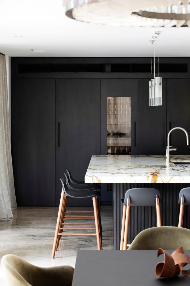 A sophisticated contemporary kitchen and dining area with full-height matte black cabinetry, integrated dark ventilation, a veined light stone island with a ribbed dark base, and a curved stainless-steel tap. Black and timber bar stools sit at the island beneath slim glass pendant lights. A partial dining table and upholstered chairs appear in the foreground, with a large sculptural ceiling light overhead. The room has pale stone flooring and a refined, high-contrast material palette.