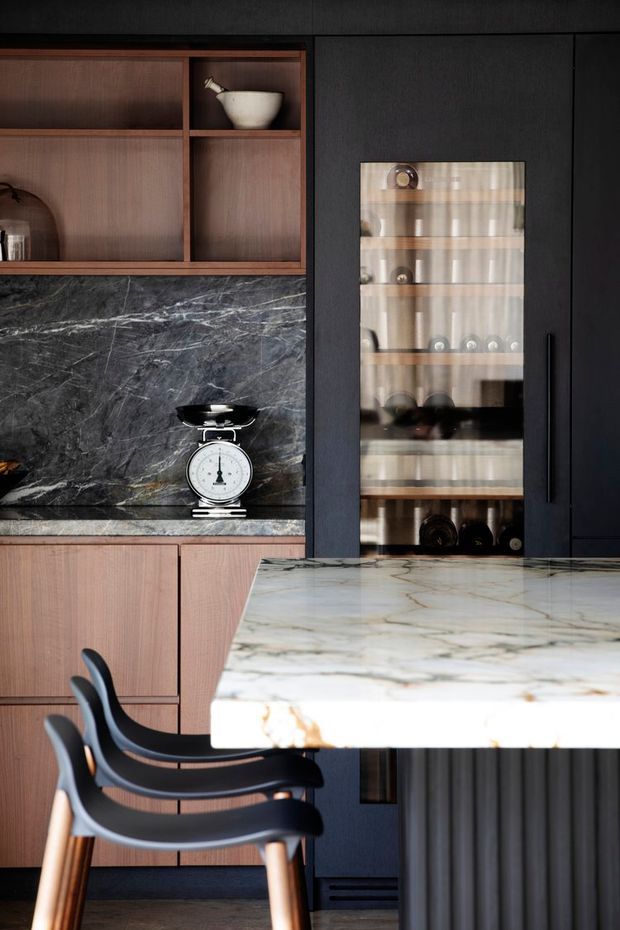 A refined, dark-toned kitchen with walnut-toned open shelving and cabinetry, dramatic veined dark stone backsplash, black panelled joinery, and a glass-front display cabinet. A pale veined stone island with a dark fluted base occupies the foreground, paired with sculptural black dining chairs. The styling is restrained, luxurious, and contemporary.