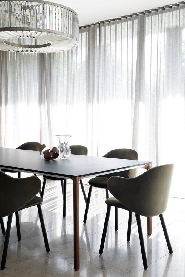 Bright, refined dining room with a rectangular dark dining table surrounded by upholstered armchairs. Floor-to-ceiling sheer curtains create a softly illuminated backdrop, while a large circular sculptural pendant light hangs above. The room features a polished light stone or tile floor and a restrained neutral palette with dark grey, white, and warm wood accents.
