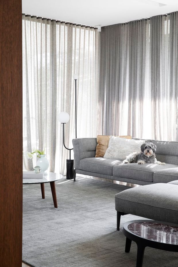 A bright, refined living room with floor-to-ceiling sheer grey curtains wrapping around corner glazing, a large grey upholstered sofa and chaise, a grey carpet, and dark wood and black accent furniture. A sculptural floor lamp and marble-topped round coffee table add understated contemporary detail. A small dog is resting on the sofa.