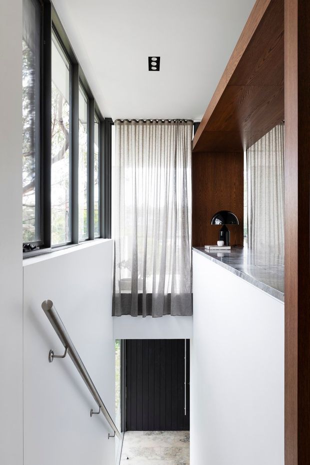 A tall, narrow contemporary interior stairwell or entry passage with a split-level arrangement. Full-height black-framed glazing lines the left wall, while a substantial dark timber built-in wall and shelving feature runs along the right. Sheer curtains soften the upper window, and a minimalist metal handrail descends beside white walls toward a black door and pale concrete floor below. The space is bright, refined, and vertically oriented.