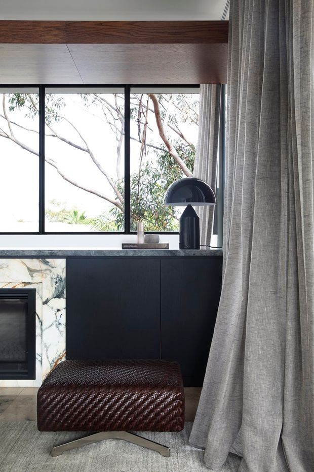 A refined living room vignette featuring a black built-in cabinet and dark stone or marble fireplace surround beneath a large black-framed window. A dark woven leather ottoman sits on a light rug, while a black dome table lamp, books and pottery decorate the cabinet. Full-height sheer grey curtains frame views of trees outside, and a timber beam adds warmth near the ceiling.