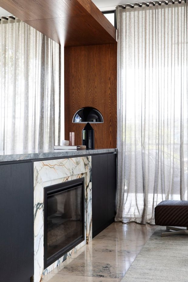 A refined living room corner featuring a built-in fireplace with a dramatic veined marble surround, dark cabinetry, rich timber wall panelling and ceiling soffit, sheer floor-to-ceiling curtains, and polished stone flooring. A black dome lamp and low upholstered lounge chair add mid-century-inspired furnishings to the elegant interior.