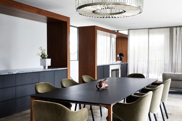 Refined contemporary dining area with a large dark rectangular dining table surrounded by upholstered olive-grey chairs. Dark built-in cabinetry with a light stone benchtop runs along the left wall, while timber-framed architectural screens divide the dining area from the living space. Floor-to-ceiling sheer curtains provide abundant filtered daylight, and a sculptural circular pendant light hangs above the table.