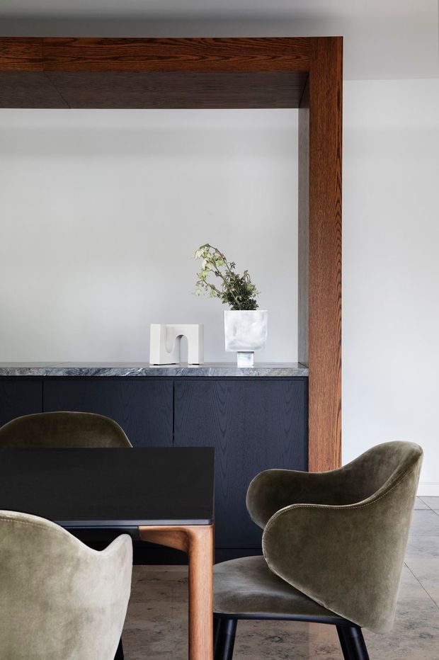 Refined dining area featuring a dark rectangular dining table surrounded by sculptural upholstered armchairs. A deep navy built-in sideboard with a grey stone benchtop runs along the rear wall, framed by a prominent dark timber architectural surround. Minimal white walls, a simple sculptural vase, and restrained contemporary styling create a sophisticated, moody interior.