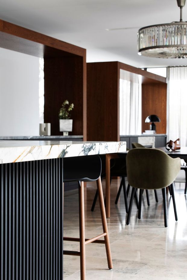 Refined open-plan interior featuring a marble-topped kitchen island with black vertically fluted cabinetry, timber-framed architectural screens, a dining table with upholstered chairs, sheer window coverings, and a statement crystal pendant light. The palette combines dark timber, black, white, and warm neutral tones with polished stone flooring.