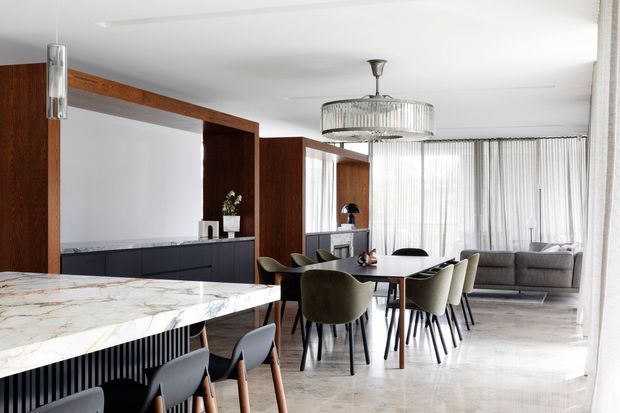 Open-plan contemporary dining and living area with a marble island in the foreground, dark timber wall panelling and cabinetry, a round dining table with upholstered chairs, a grey sofa, and full-height sheer curtains. A sculptural pendant light hangs above the dining table, while the bright neutral palette and refined materials create an elegant, spacious interior.