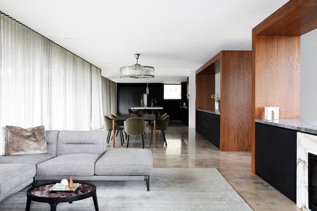 Open-plan contemporary interior combining a living room, dining area and kitchen. A large light-grey sectional sofa sits beside a black round coffee table, while sheer floor-to-ceiling curtains line the left wall. The dining zone includes a dark table with upholstered chairs and a decorative pendant light. At the rear and right side, the kitchen features dark cabinetry, rich walnut-toned timber joinery, a stone island benchtop and a black splashback or wall finish. The space has polished stone or concrete-look flooring, white ceilings, and a refined neutral palette with strong black and timber accents.