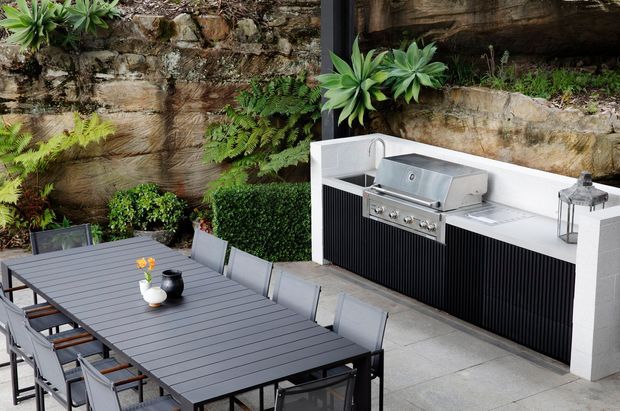 A landscaped courtyard terrace with a large outdoor dining table and chairs beside a built-in outdoor kitchen. The kitchen features a stainless-steel barbecue, sink, white counter and side walls, and dark vertical-ribbed cabinetry. Stone retaining walls, timber screening, and lush ferns and succulents create a secluded garden setting.