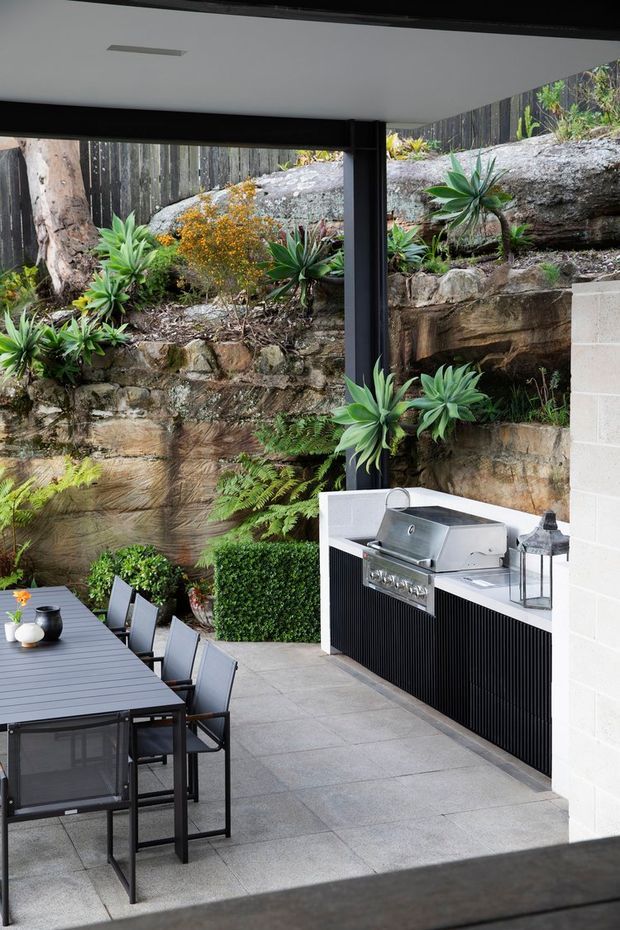 A contemporary covered outdoor entertaining area with a built-in stainless-steel barbecue and black vertically ribbed cabinetry, positioned beside a long outdoor dining table. Large grey paving slabs, dark structural framing and a white ceiling define the space, while layered stone retaining walls and lush planting create a landscaped backdrop.