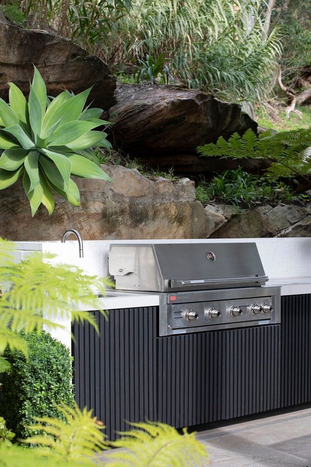 A landscaped outdoor kitchen set against a lush, natural rock garden. The built-in kitchen features a stainless-steel barbecue, sink, light benchtop, and dark vertical-panel cabinetry, surrounded by ferns, broad-leaf planting, stone retaining elements, and tiled paving.