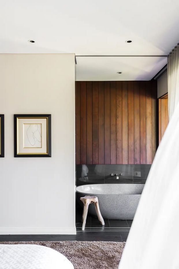 A refined bedroom interior with a partially enclosed ensuite bathroom. The bathroom features a freestanding textured grey bathtub, dark grey tiled floor and wall base, a glossy dark stone or tile ledge, and a warm vertical timber feature wall. The bedroom has soft white walls, recessed ceiling lights, framed artwork, plush neutral carpeting, and sheer curtains beside a window. The composition looks toward the bathroom from the bedroom, creating a calm, warm, contemporary retreat.