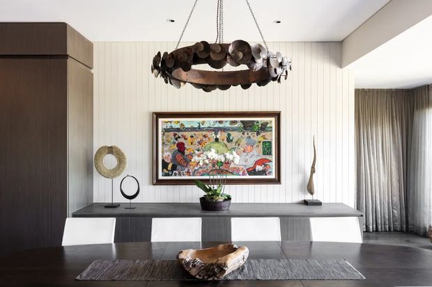 A refined dining room with a long dark timber dining table and contemporary white chairs. The room features vertical light wall panelling, dark built-in cabinetry, a large framed colourful artwork, sculptural accessories, and a distinctive circular pendant light made from layered metal forms. Full-height curtains open toward an adjacent living area, creating a sophisticated, textural interior.