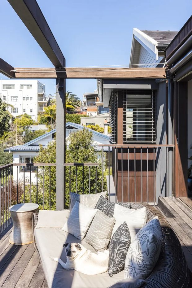 A bright elevated outdoor terrace or balcony with dark timber decking, a cushioned outdoor sofa, and a timber pergola structure. The space overlooks neighbouring homes and greenery, with a modern dark-clad dwelling, horizontal window louvres, and metal balustrading visible. Strong sunlight and clear blue sky create an airy residential setting.