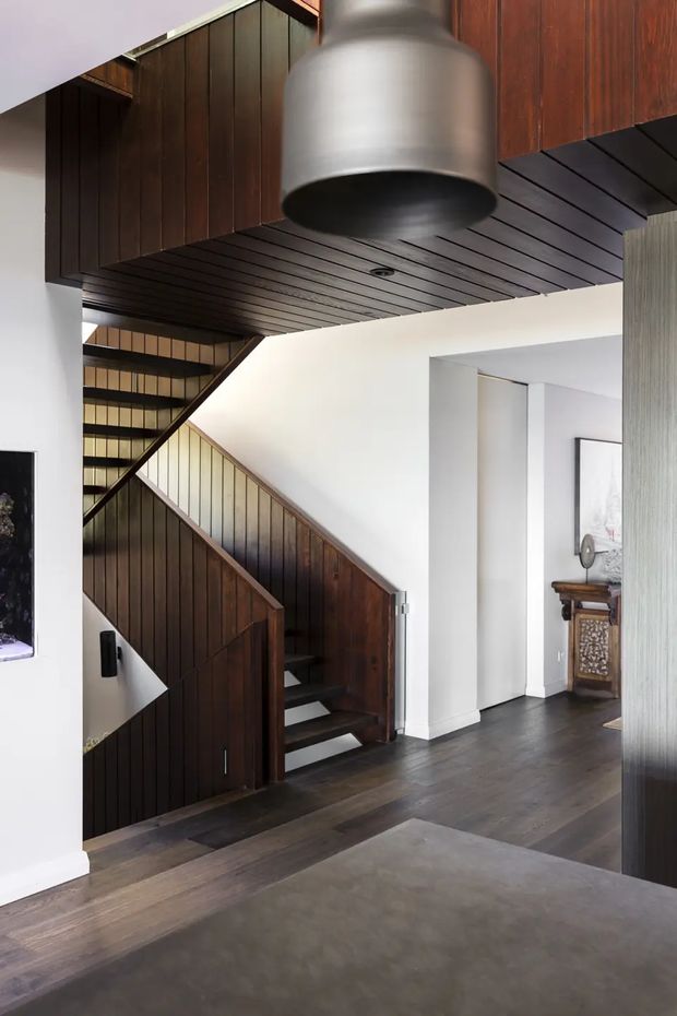 Interior view of a contemporary entry and stair hall featuring a sculptural dark timber staircase with matching vertical wall panelling and a slatted timber ceiling. Dark timber flooring, crisp white walls, a large brushed-metal pendant light, and a glimpse of console furniture in the adjacent room create a refined, dramatic composition.