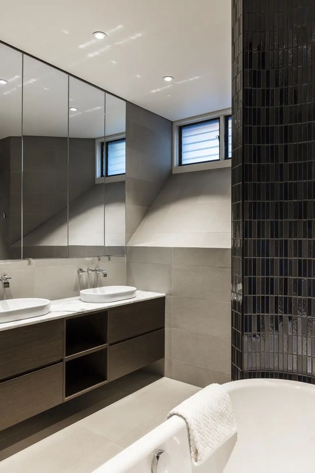 Contemporary bathroom with a double vanity, large mirrored wall, pale grey tiled walls and floor, and dark wood floating cabinetry. A freestanding white bathtub occupies the foreground, while a tall feature wall clad in glossy narrow dark tiles creates a strong vertical accent. High horizontal windows and recessed ceiling lights provide natural and artificial illumination.
