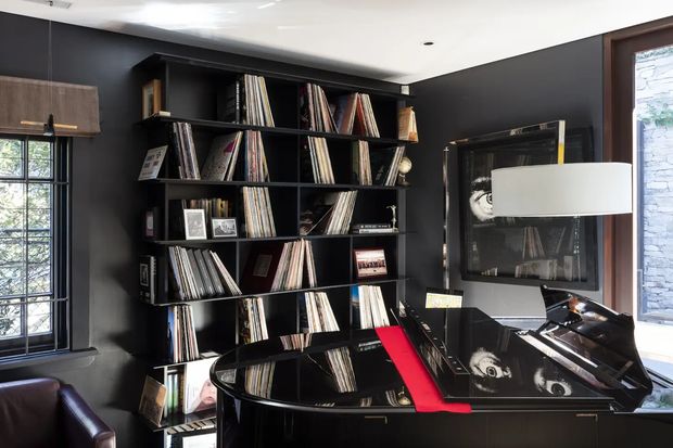 A dark, sophisticated music room centred on a glossy black grand piano with a vivid red runner. A large black built-in shelving unit displays vinyl records and books, while black walls, framed artwork, tall multi-pane windows, and a contemporary floor lamp complete the dramatic interior.