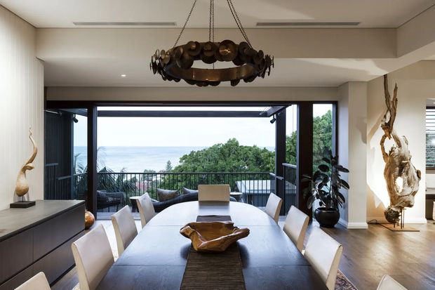 A refined dining room with a long dark dining table, upholstered dining chairs, sculptural timber centerpiece, and a striking circular metal pendant light. Full-height dark-framed glazing opens to a balcony with an ocean view, while sculptural wood artwork and decorative objects add warmth to the neutral interior.