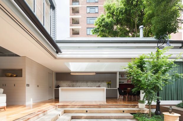 Bright contemporary inner-city courtyard renovation with an open-plan kitchen and dining area opening onto stepped outdoor paving. The kitchen features pale built-in cabinetry, a marble-look island, timber flooring, skylights, and large glazed openings. A leafy courtyard with mature planting and a black garden lamp occupies the foreground, while an apartment building is visible beyond the roofline.