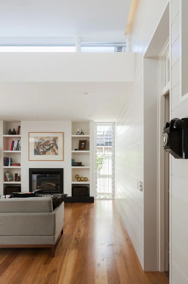 Bright, narrow living room viewed from an adjacent hallway or entry, with warm timber flooring, white-painted horizontal wall panelling, built-in open shelving, a black fireplace surround, a neutral sofa, and a high clerestory-style opening that brings in daylight. A glazed door or tall window at the rear adds further light, while a black wall-mounted object is visible near the right edge.