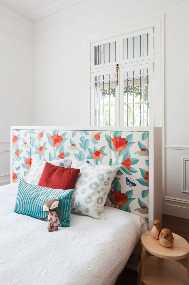 Bright, playful bedroom corner featuring a bed with a vivid floral upholstered headboard, layered patterned cushions, a turquoise bolster, and a small wooden bedside table with decorative wooden birds. The room has white walls, decorative wall panelling and cornices, dark timber flooring, and a tall white-framed leadlight-style window with security bars. The overall interior is cheerful and eclectic with strong botanical colours.