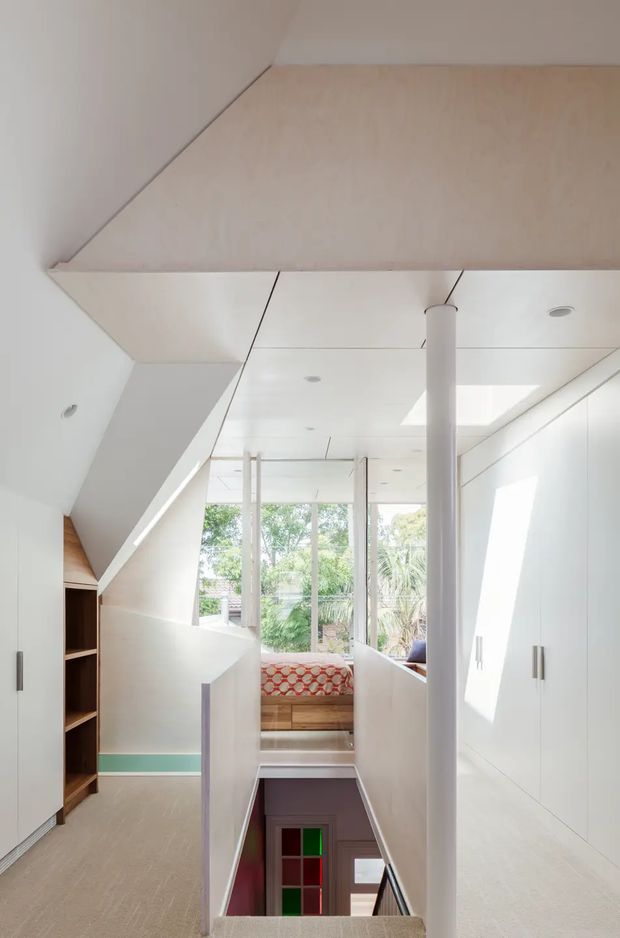 Bright, compact modern interior hallway and stair landing with white built-in cabinetry, pale timber flooring, sloped ceiling planes, slender white structural columns, and a skylight. The view looks toward a sunlit window seat overlooking greenery, with a stair opening descending to a lower level and a glimpse of a colourful room below.