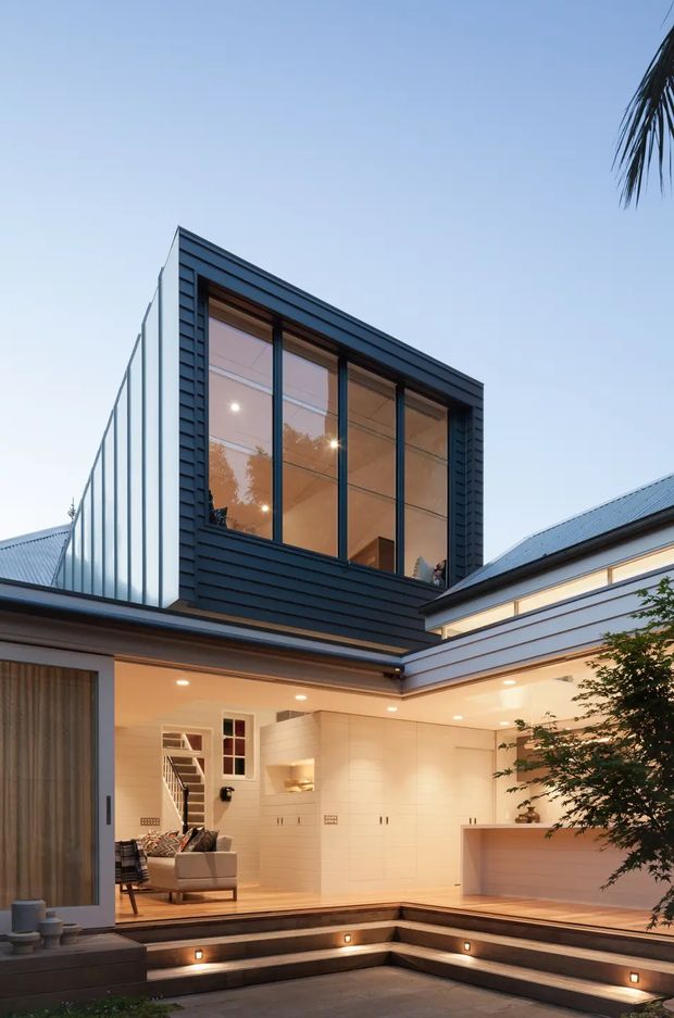 Twilight view of a contemporary multi-level courtyard home with dark horizontal cladding, expansive black-framed glazing, and a warm, sheltered outdoor living area. The courtyard includes stepped timber decking and built-in illuminated seating or steps, while the open ground floor reveals a living room and a minimalist kitchen with pale cabinetry and a peninsula. The architecture uses strong geometric forms, a projecting upper-level glazed volume, and a mix of dark and light facade finishes.
