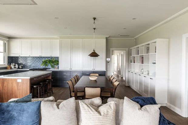 Open-plan interior viewed from the living area, featuring a large white shaker-style kitchen with dark lower cabinetry, a dark stone island, tiled backsplash, and timber cabinetry panels. A dark rectangular dining table with upholstered chairs sits centrally, while built-in white shelving and storage line the right wall. The foreground includes a large light sofa with blue cushions and throws. The room is bright, refined, and casually coastal in character.