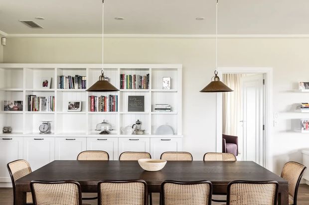 Bright, refined dining room with a large dark timber dining table surrounded by woven cane-back chairs. A full wall of white built-in cabinetry and open bookshelves provides storage and display, while two bronze pendant lights hang above the table. Pale neutral walls, white trim, mirrored panels, and a doorway to an adjacent room create an airy, classic interior.