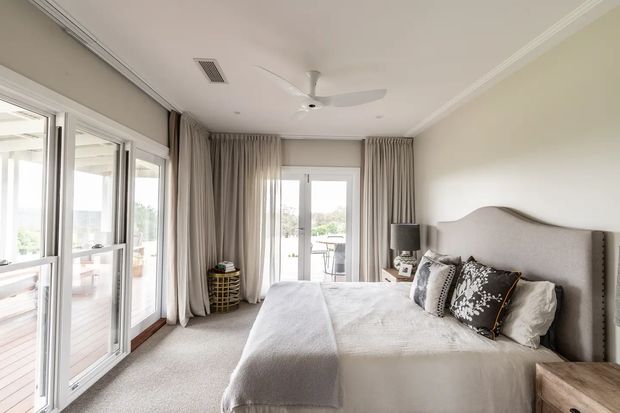 Bright, softly styled bedroom with a large upholstered bed, layered neutral bedding and decorative cushions. Full-height glazed doors and windows open toward a balcony or outdoor area, with floor-length taupe curtains, pale carpet, white walls, a ceiling fan and simple bedside furniture. The room has a calm, refined contemporary-traditional feel.