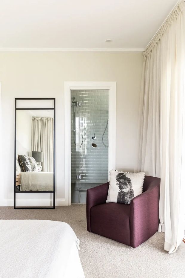 A bright, refined bedroom with pale cream walls, carpeted flooring, full-height sheer curtains, and a large black-framed leaning mirror reflecting the bed. A burgundy upholstered armchair with a bird-print cushion sits beside the window. An open ensuite shower is visible directly from the bedroom, featuring pale green subway wall tiles and chrome fixtures. Traditional trim and restrained furnishings create a calm, elegant atmosphere.