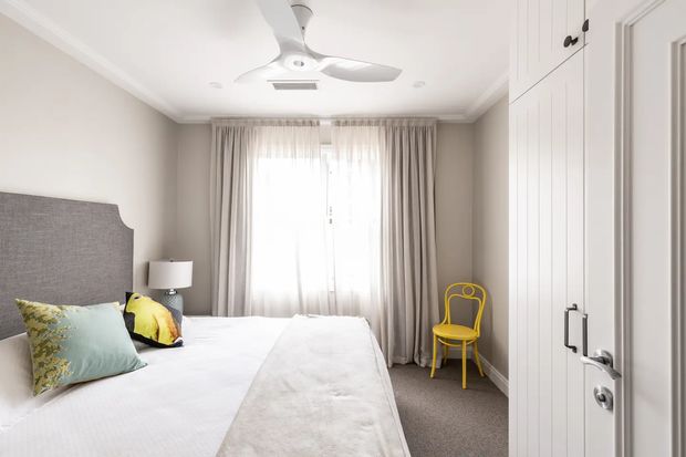 Bright, compact bedroom viewed from the doorway, with a large upholstered grey headboard, white bedding, bedside lamp, full-height curtains over a window, built-in white panelled wardrobe cabinetry, and a distinctive yellow accent chair. A white ceiling fan and neutral carpet complete the simple, softly styled interior.
