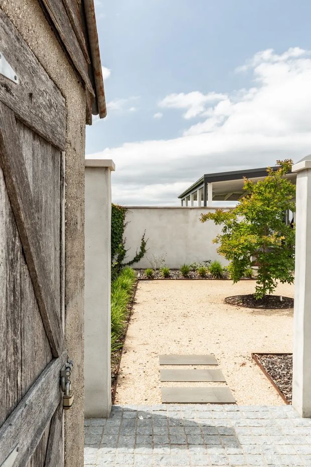 View through an open rustic timber gate into a minimalist landscaped courtyard. Pale rendered boundary walls frame a light gravel garden with rectangular stepping stones, dark planted beds, clipped greenery, and a small tree. A contemporary flat-roofed structure is visible beyond the courtyard. The foreground has blue-grey stone paving, while the image is brightly lit under a partly cloudy sky.