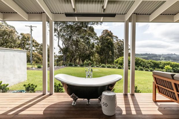 Open-air covered outdoor bathing area with a freestanding clawfoot bathtub positioned on a timber deck. The space has a corrugated metal ceiling, pale structural posts, a dark ceiling fan or light fixture, and views across a manicured lawn and rural landscape. Outdoor seating is visible at the right.