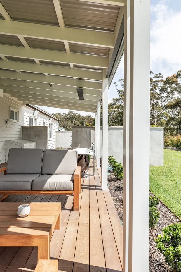 A bright covered outdoor deck beside a light-clad house, featuring a timber deck, timber-framed grey outdoor sofa, low coffee table, dining setting, white structural posts, and a corrugated translucent roof supported by exposed beams. The deck overlooks a landscaped lawn and garden with mature trees beyond.