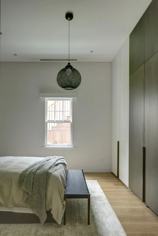 A calm, contemporary bedroom with a partially visible neatly made bed, soft grey textured throw, pale rug, light timber flooring, and a dark woven bench. Full-height built-in wardrobes line the right wall, combining white panels with dark timber doors and slim vertical handles. A woven spherical pendant hangs centrally from the smooth white ceiling, while a small window brings in natural light.