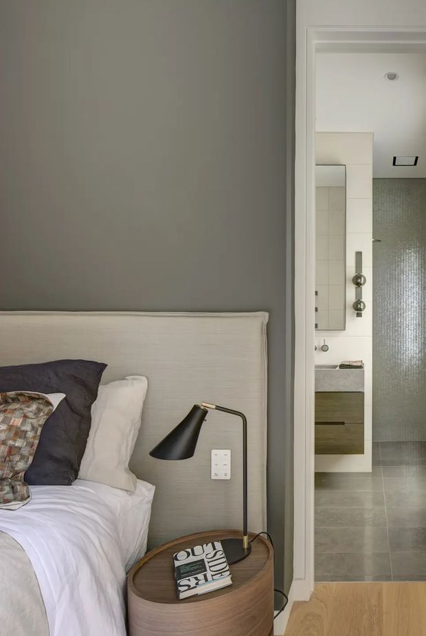 View from a refined bedroom into an adjacent bathroom. The bedroom features a softly upholstered light headboard, layered white and charcoal bedding, a black angled bedside lamp, and a rounded timber bedside table. The bathroom beyond has pale tile surfaces, a wall-mounted vanity with timber-toned drawers, a large mirror, and a textured glass shower enclosure. The overall palette is muted, neutral, and contemporary.