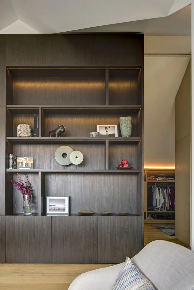 Interior view of a refined bedroom with a full-height dark timber built-in shelving and display unit, integrated warm lighting, and an open walk-in wardrobe behind it. The lower right foreground shows part of a light upholstered bed with patterned cushions. The shelving contains books, ceramics, framed artwork, sculptures, and decorative objects. Light timber flooring, a neutral ceiling, and concealed lighting create a warm, sophisticated atmosphere.