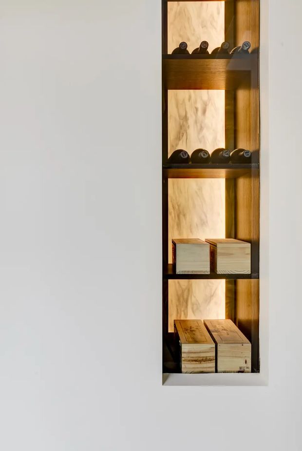 A narrow built-in display niche set into a white wall, with dark timber shelving and a warm wood-grain backing. The shelves display horizontally stored bottles and small wooden boxes, illuminated by warm integrated lighting. The composition is minimal and focused on the custom joinery.