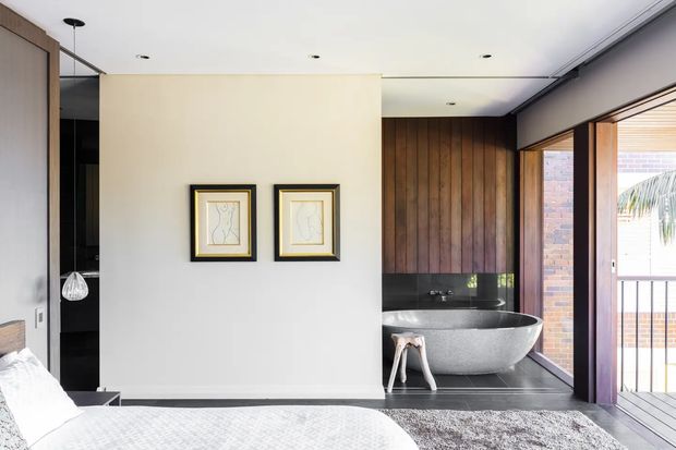 A bright contemporary bedroom with a partially visible bed in the foreground and an open ensuite bathroom beyond. The bathroom features a freestanding stone-look bathtub, dark tiled flooring, black fixtures, a warm vertical timber feature wall, and extensive glazing opening toward a balcony. The bedroom has pale walls, recessed downlights, framed artwork, and neutral textured furnishings.