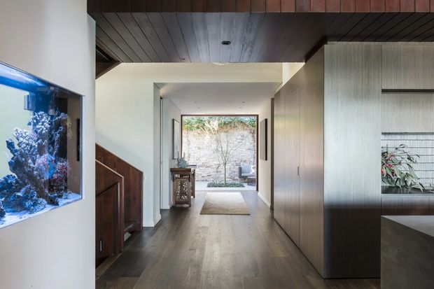 A refined residential hallway or entry passage with dark timber ceiling panels, warm timber flooring, built-in timber cabinetry, and a textured vertical-panel feature wall. The corridor opens toward a small landscaped courtyard framed by glazing or an open doorway. A large built-in aquarium is visible on the left, while dark joinery and a recessed display niche define the right side. The space has a calm, warm, contemporary architectural character with strong indoor-outdoor connection.