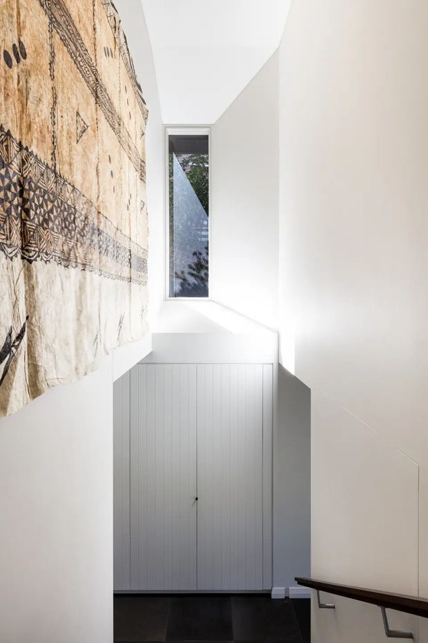 A narrow, bright stairway or landing with crisp white walls, a slim vertical window, dark flooring, and a tall built-in cupboard beneath the stairs. A large patterned textile hangs along the left wall, while a dark timber handrail with metal brackets runs down the right side. The composition emphasizes minimalist geometry, strong natural light, and high contrast between white surfaces and dark floor and railing.