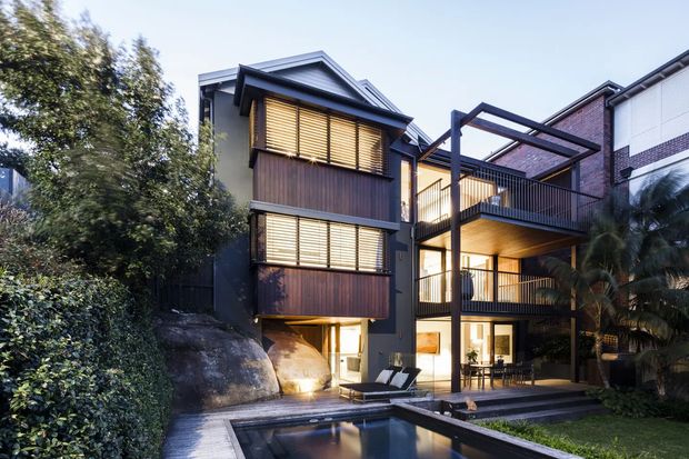 A multi-storey contemporary residential house viewed from the landscaped rear courtyard, with dark timber-clad upper-level volumes, extensive glazing, black steel-framed balconies and a covered outdoor living area. A rectangular swimming pool occupies the foreground, while large natural rock outcrops, dense planting and palm trees frame the building. The roof forms are pitched and gabled, and warm interior lighting contrasts with the dark exterior finishes.