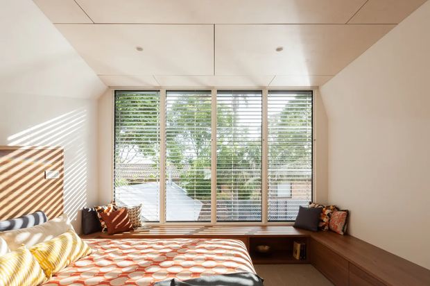 Bright compact bedroom with a built-in timber window seat and storage bench, a low bed, patterned bedding, and full-height glazing fitted with horizontal blinds. Sunlight creates strong striped shadows across the pale walls and bed, giving the room a warm, contemporary feel.