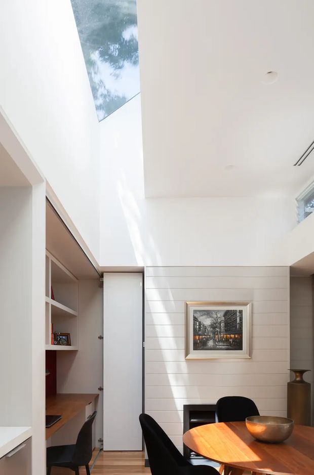 Bright contemporary dining and study area with a double-height white ceiling, skylight, built-in shelving and desk, white horizontal wall panelling, framed artwork, and a timber dining table with dark upholstered chairs. Strong natural light creates a clean, airy atmosphere.