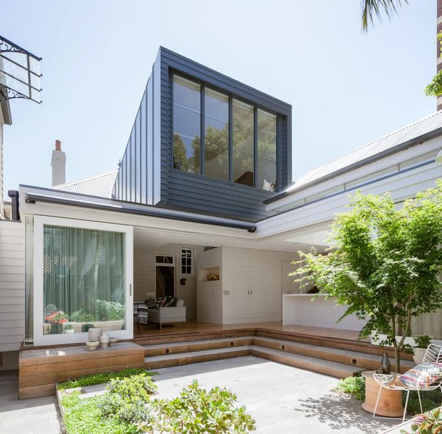 Bright architectural photograph of a contemporary courtyard extension. The central courtyard has stepped timber seating, pale paved flooring, built-in white joinery, a large glazed sliding door, and a two-storey dark-clad box with expansive windows above. White horizontal cladding and a light metal roof surround the courtyard, with integrated planting and outdoor chairs adding a residential outdoor-living feel.