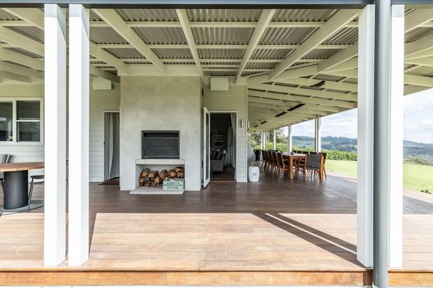 Open-air covered entertaining terrace with a large timber deck, exposed white-painted structural beams and corrugated ceiling, an outdoor dining setting, and a built-in masonry fireplace with stacked firewood. The space opens onto a broad green rural landscape and is brightly lit by daylight.