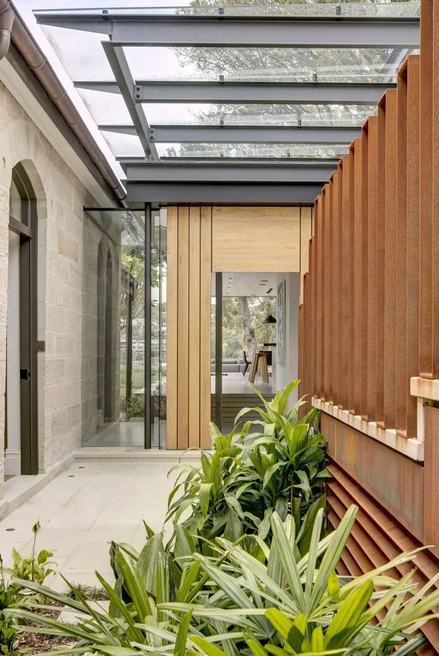 A narrow glazed architectural link or courtyard passage between an older masonry building and a contemporary extension. The passage has pale tiled or concrete paving, full-height glazing, a steel-framed transparent roof with exposed beams, light timber wall panels, and a prominent rust-toned vertical screening facade. Lush planting occupies the foreground, while an interior living space is visible through the glazed opening at the far end.
